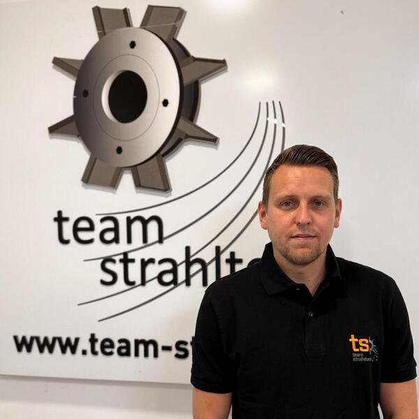 Mann in schwarzem Shirt vor Logo von Team Strahltechnik, neutraler Ausdruck.