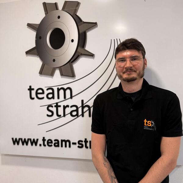 Person mit Brille lächelt leicht neben dem Team-Logo, trägt ein schwarzes Polo-Shirt.
