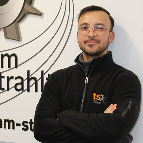 Ein lächelnder Mann mit Brille und Bart steht mit verschränkten Armen vor einem Logo