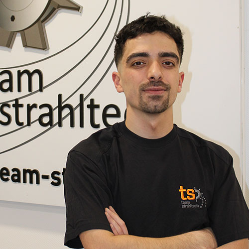 Junger Mann mit dunklem Haar und dunklem Schnurrbart lächelt selbstbewusst vor einem weißen Team-Strahltechnik Schild in schwarzem Shirt